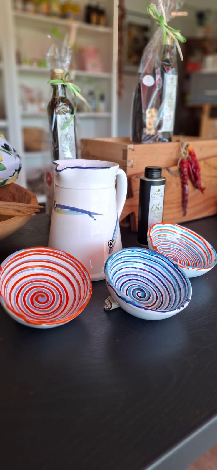 White ceramic pitcher with blue trim and three spiral-pattern bowls (red and blue) on a dark table, with gift-wrapped bottles and a wooden crate in the background.