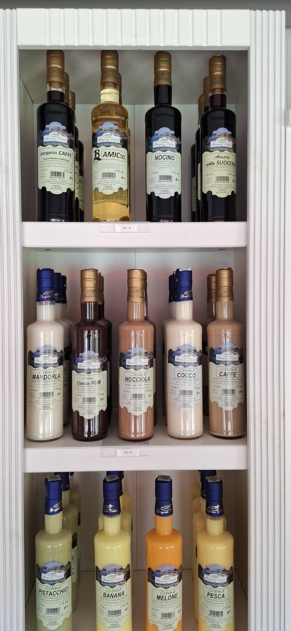 Row of Italian crema liqueur bottles on white shelves, flavors like mandorla, nocciola, cocco, pistacchio, banana, melone, pesca visible in labels.
